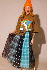 Model wearing a brown graphic shirt with a colorful graphic design of a rainbow and unicorn, an orange beanie, and a long, patterned skirt, with sections of black and white polka dots, blue flannel, brown flannel, and leopard prints, against a plain background, frontal view