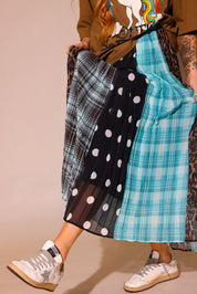 Model wearing a brown graphic shirt with a colorful graphic design of a rainbow and unicorn, an orange beanie, and a long, patterned skirt, with sections of black and white polka dots, blue flannel, brown flannel, and leopard prints, against a plain background, lower close up frontal view