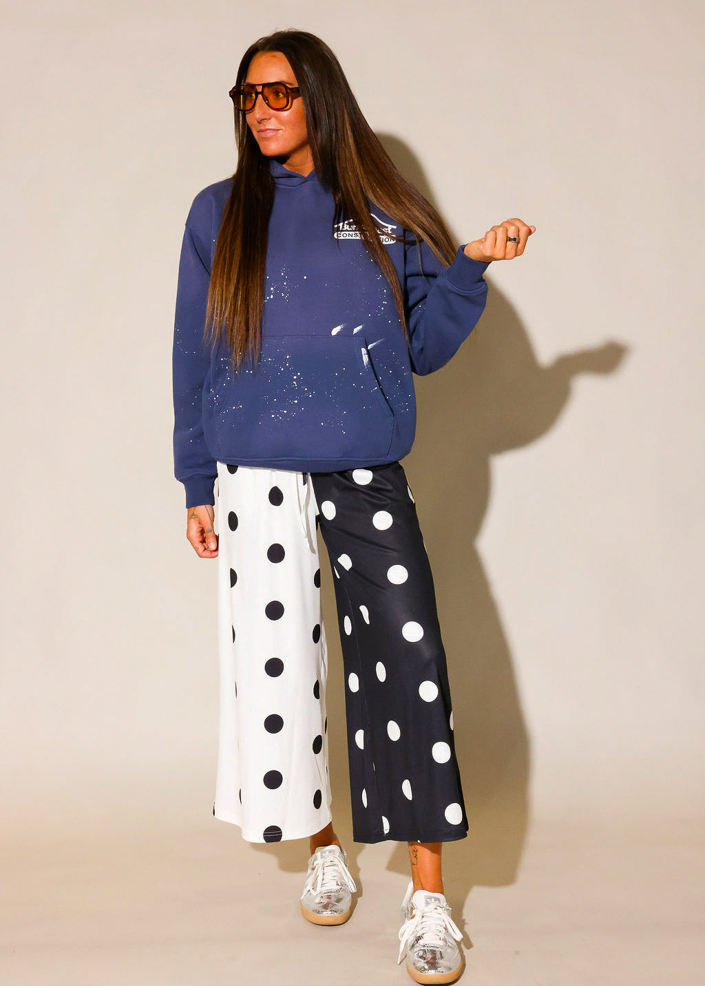 Model wearing a blue hoodie with white paint splashes and white burnt toast construction logo on left side and black and white polka dot pants  and sunglasses, against a beige background, frontal view