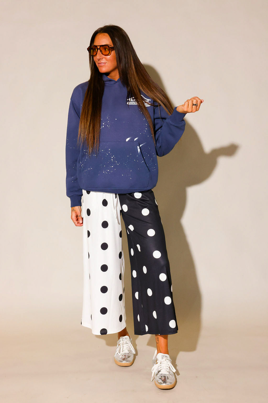 Model wearing a blue hoodie with white paint splashes and white burnt toast construction logo on left side and black and white polka dot pants  and sunglasses, against a beige background, frontal view