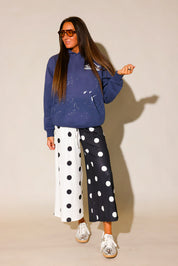 Model wearing a blue hoodie with white paint splashes and white burnt toast construction logo on left side and black and white polka dot pants  and sunglasses, against a beige background, frontal view