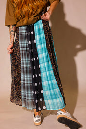 Model wearing brown graphic tee and a long, patterned skirt, with sections of black and white polka dots, blue flannel, brown flannel, and leopard prints, show the back slip underneath, against a plain background, lower half of body close up back view