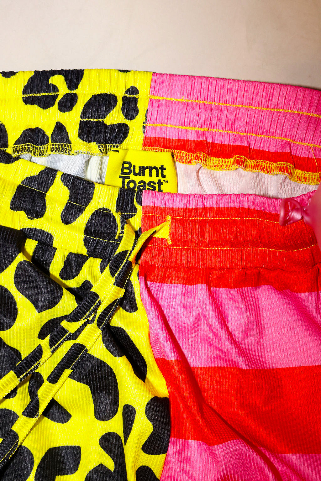 Colorful half yellow leopard print pants, and half pink striped pants, with a burnt toast label in yellow, on a plain background, flat lay close up frontal view