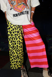 Model wearing a white short sleeve shirt with a tiger graphic and text, half yellow leopard print pants, and half pink striped pants, with hands in pockets, lower half of body frontal view