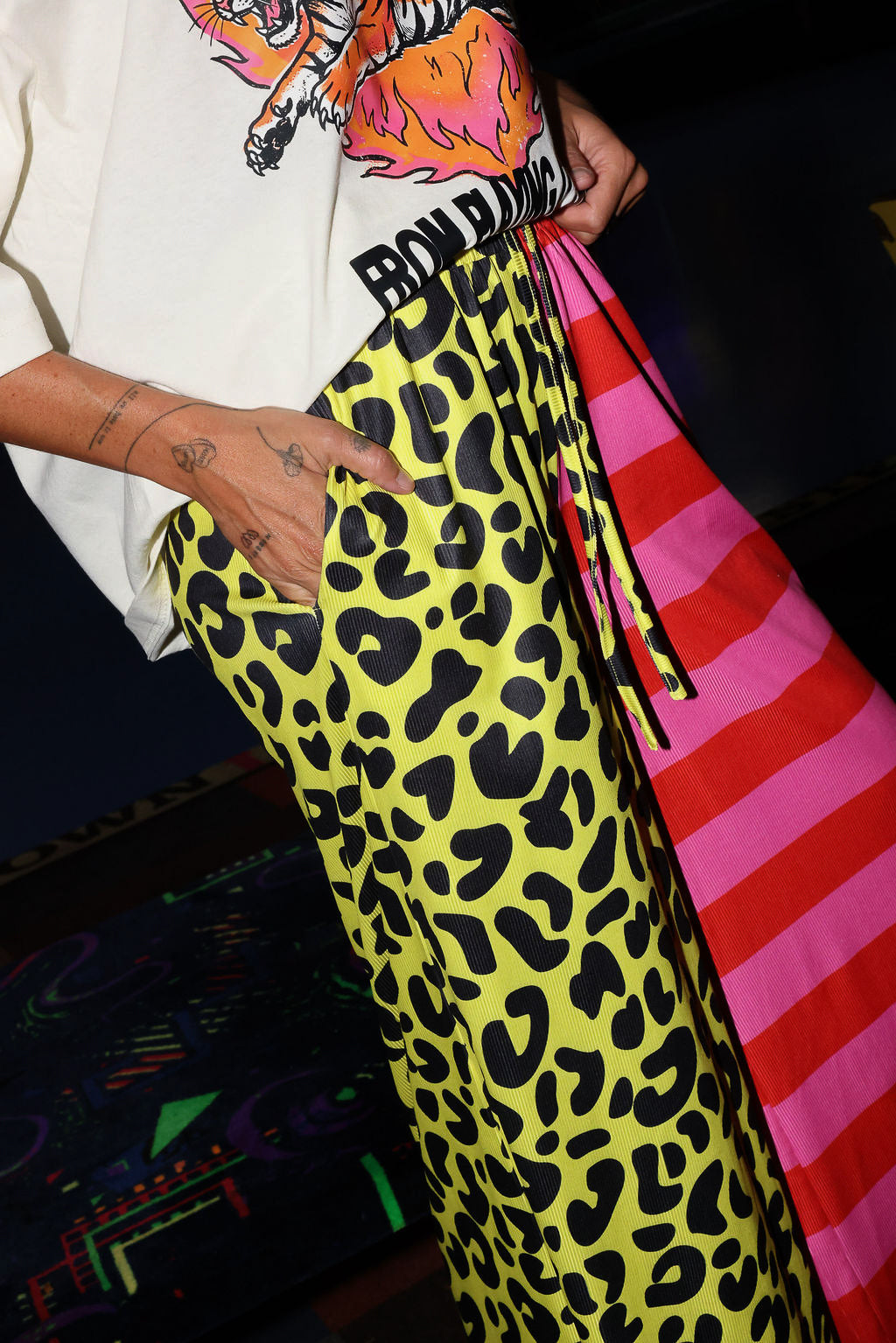 Model wearing a white short sleeve shirt with a tiger graphic and text, half yellow leopard print pants, and half pink striped pants, with hands in pockets, lower half of body side view