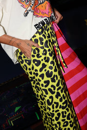 Model wearing a white short sleeve shirt with a tiger graphic and text, half yellow leopard print pants, and half pink striped pants, with hands in pockets, lower half of body side view