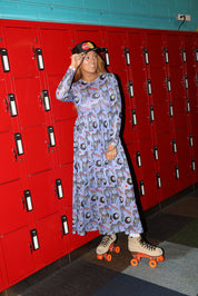 Model wearing a black trucker hat with red and yellow circle logo, long purple mesh dress with a zebra, rainbow and 8 ball design print, and roller skates, in front of red lockers, full frontal view