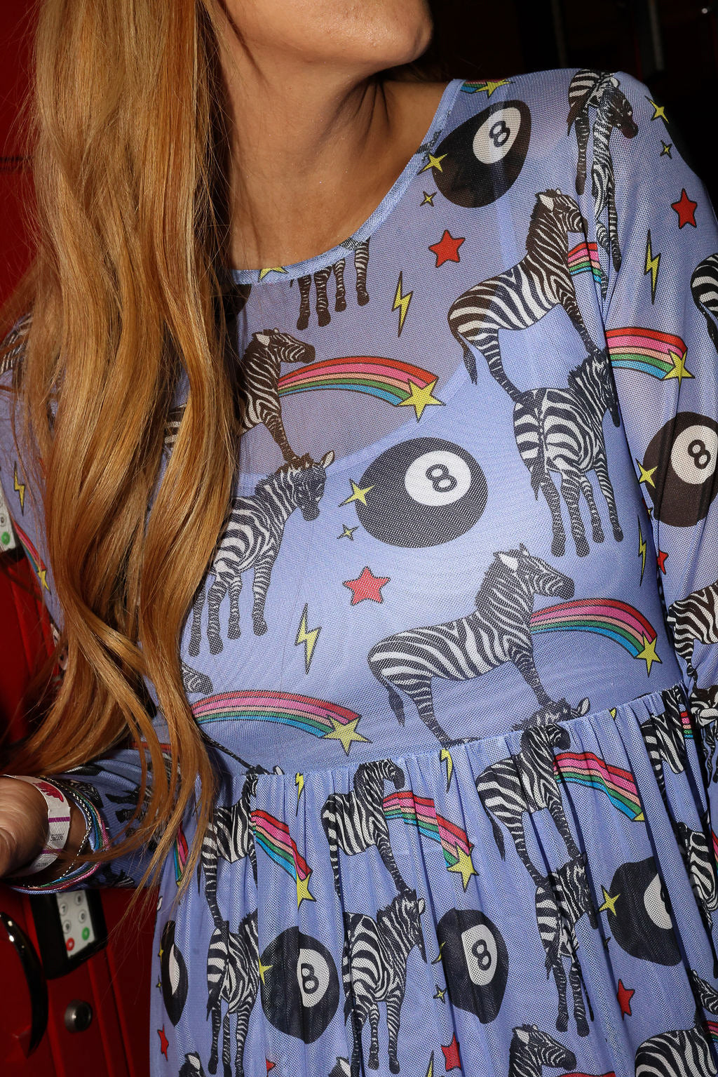 Model wearing a long purple mesh dress with a zebra, rainbow and 8 ball design print, in front of red lockers, lose up frontal view