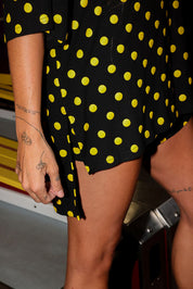 Model wearing a black and yellow polka dot set with a short sleeve top with a slide on the side, and biker shorts, standing in front of a vintage arcade machine in an indoor setting, close up side view