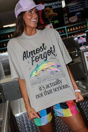A woman wearing a light gray graphic t-shirt with a colorful dolphin illustration and the text 'Almost Forgot! it's actually not that serious.' She is also wearing a lavender baseball cap and multicolored striped shorts, standing in a cafe setting