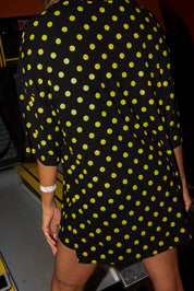 Model wearing a black and yellow polka dot set with a short sleeve top and biker shorts, standing in front of a vintage arcade machine in an indoor setting, close up back view