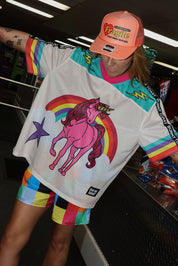 Model wearing a neon orange trucker hat that reads Fragile and has a red heart, colorful mesh short sleeve jersey with a unicorn and rainbow design, rainbow checkered pattern shorts, standing in an indoor setting. close up side view