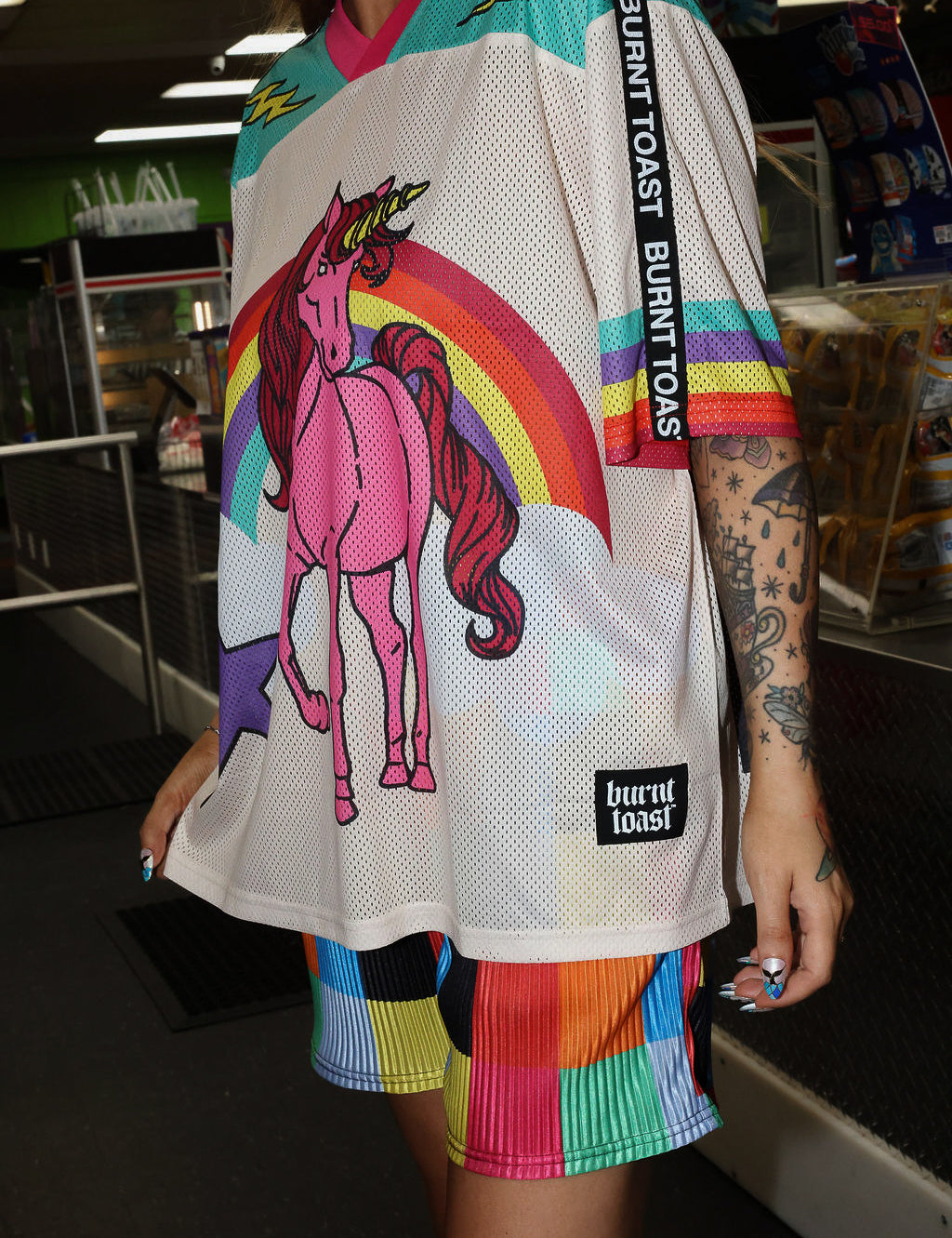 Model wearing a colorful mesh short sleeve jersey with a unicorn and rainbow design, rainbow checkered pattern shorts, standing in an indoor setting. close up frontal view