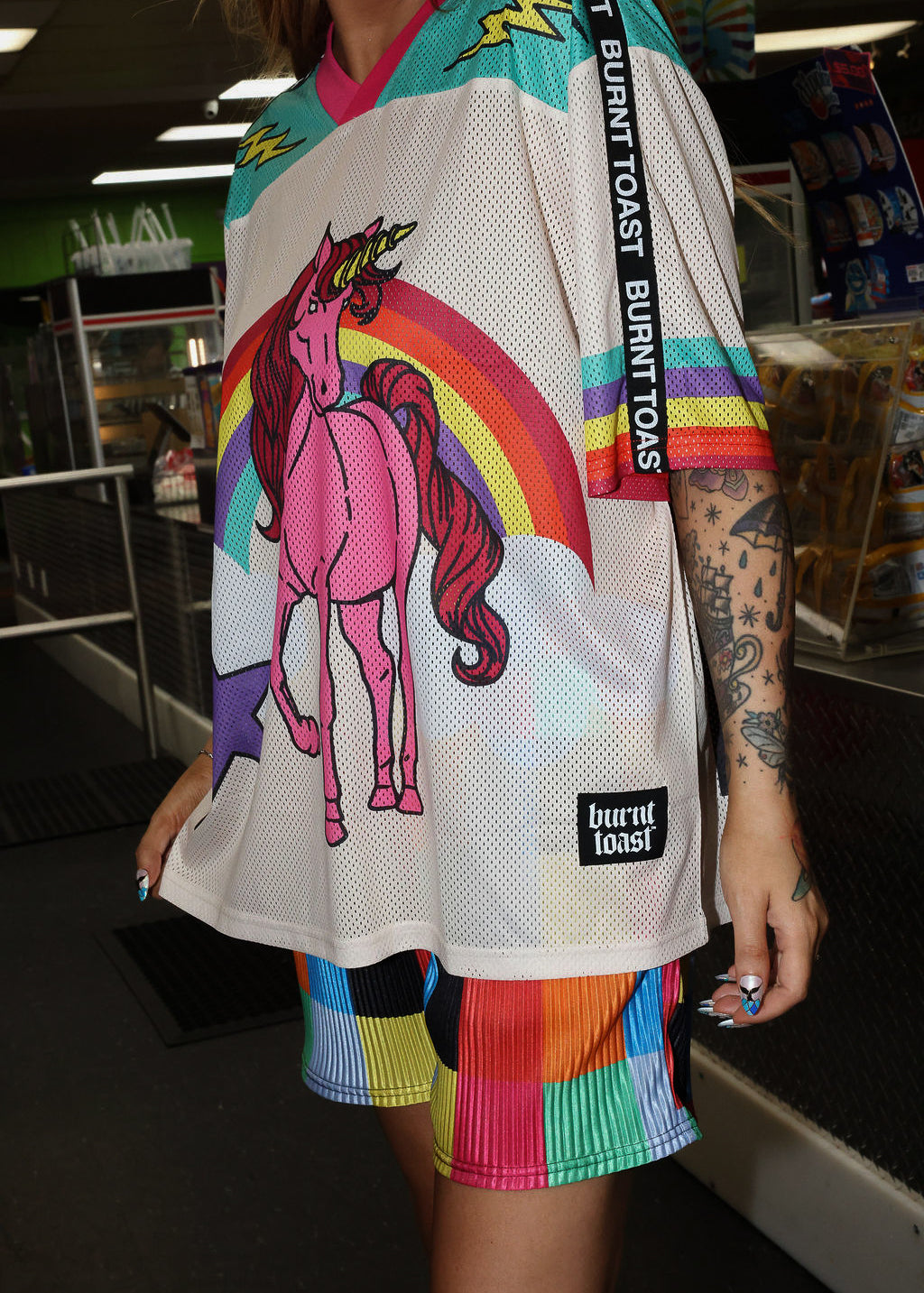 Model wearing a colorful mesh short sleeve jersey with a unicorn and rainbow design, rainbow checkered pattern shorts, standing in an indoor setting. close up frontal view