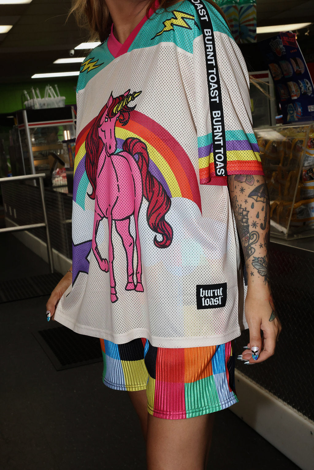 Model wearing a colorful mesh short sleeve jersey with a unicorn and rainbow design, rainbow checkered pattern shorts, standing in an indoor setting. close up frontal view