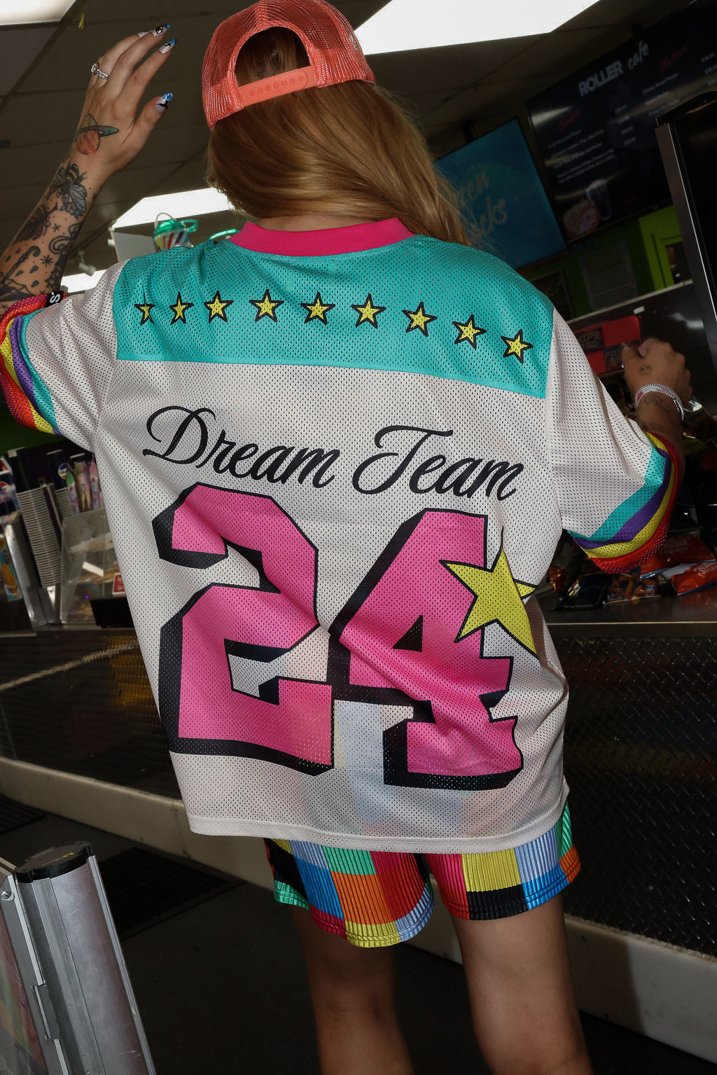 Model wearing a neon orange trucker hat, colorful mesh short sleeve jersey with the number 24 , stars, and 'Dream Team' text on the back, rainbow checkered pattern shorts, standing in an indoor setting. close up back view