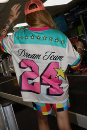 Model wearing a neon orange trucker hat, colorful mesh short sleeve jersey with the number 24 , stars, and 'Dream Team' text on the back, rainbow checkered pattern shorts, standing in an indoor setting. close up back view