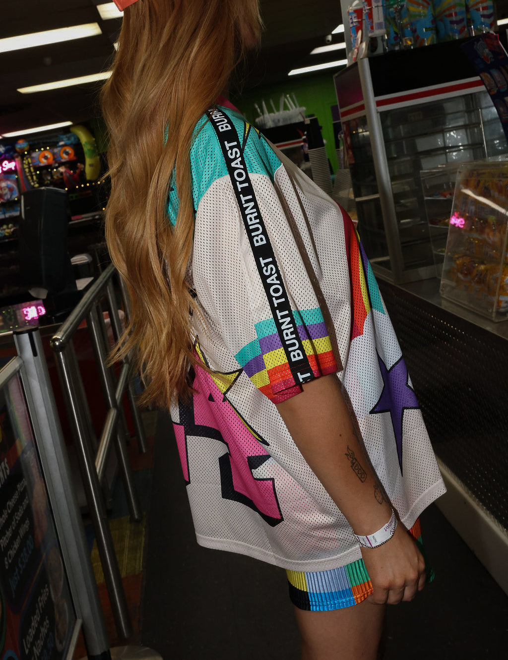Model wearing a neon orange trucker hat, colorful mesh short sleeve jersey with a unicorn and rainbow design, rainbow checkered pattern shorts, standing in an indoor setting. close up side view