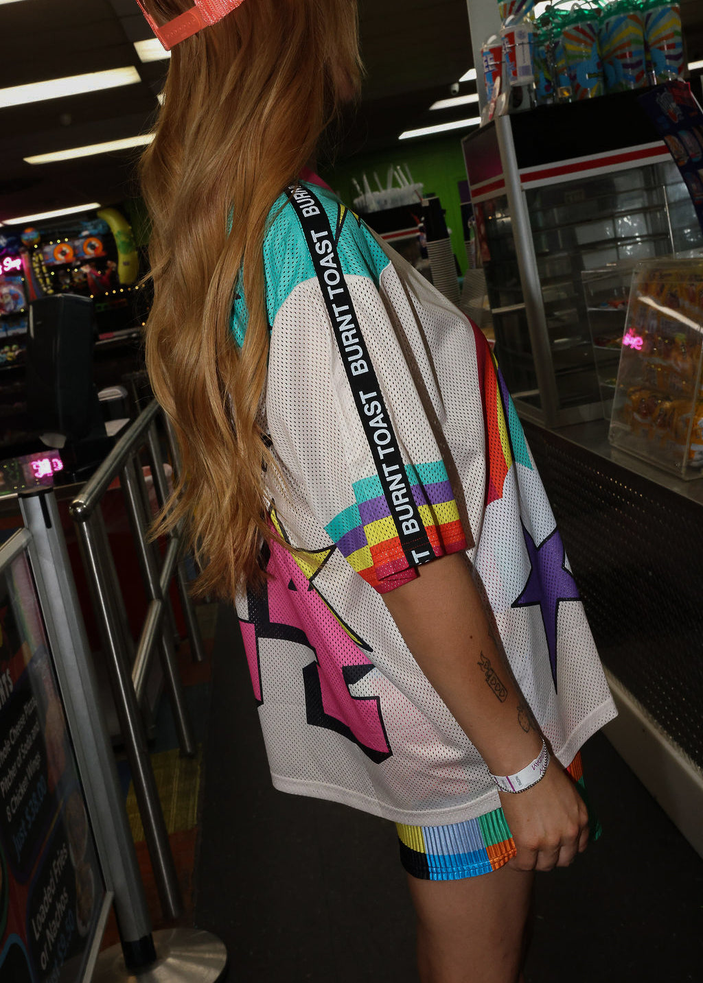 Model wearing a neon orange trucker hat, colorful mesh short sleeve jersey with a unicorn and rainbow design, rainbow checkered pattern shorts, standing in an indoor setting. close up side view