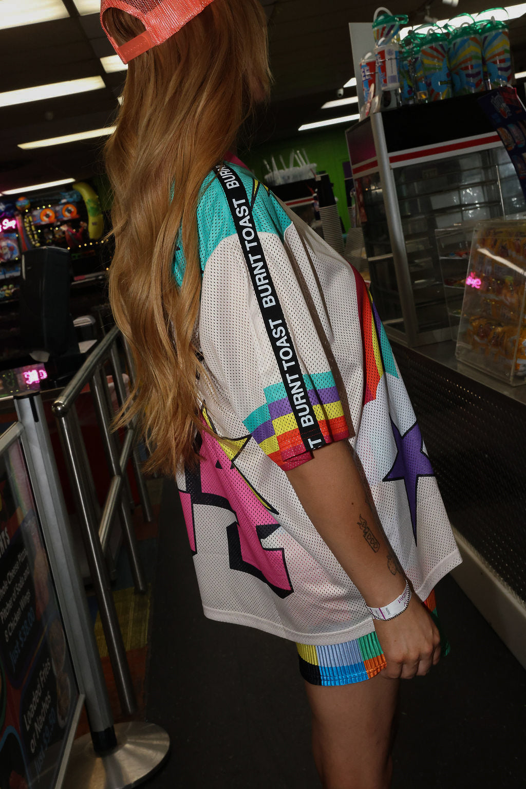 Model wearing a neon orange trucker hat, colorful mesh short sleeve jersey with a unicorn and rainbow design, rainbow checkered pattern shorts, standing in an indoor setting. close up side view