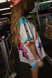 Model wearing a neon orange trucker hat, colorful mesh short sleeve jersey with a unicorn and rainbow design, rainbow checkered pattern shorts, standing in an indoor setting. close up side view