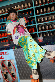 Model wearing a black beanie, colorful mesh short sleeve jersey with a long sleeve mesh top with color polka dots, satin seafoam green pants with yellow smiley faces, sitting on a desk with roller skates in the background, frontal view