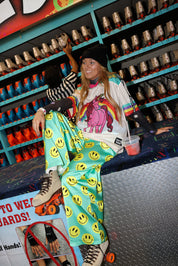 Model wearing a black beanie, colorful short sleeve jersey with long sleeve black mesh top, sitting on a desk, with colorful seafoam green pants with yellow smiley faces, in a store setting, frontal view