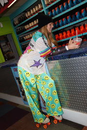 Model wearing a black beanie, colorful short sleeve jersey with long sleeve black mesh top, standing next to a desk, with colorful seafoam green pants with yellow smiley faces, and roller skates, in a store setting, frontal view