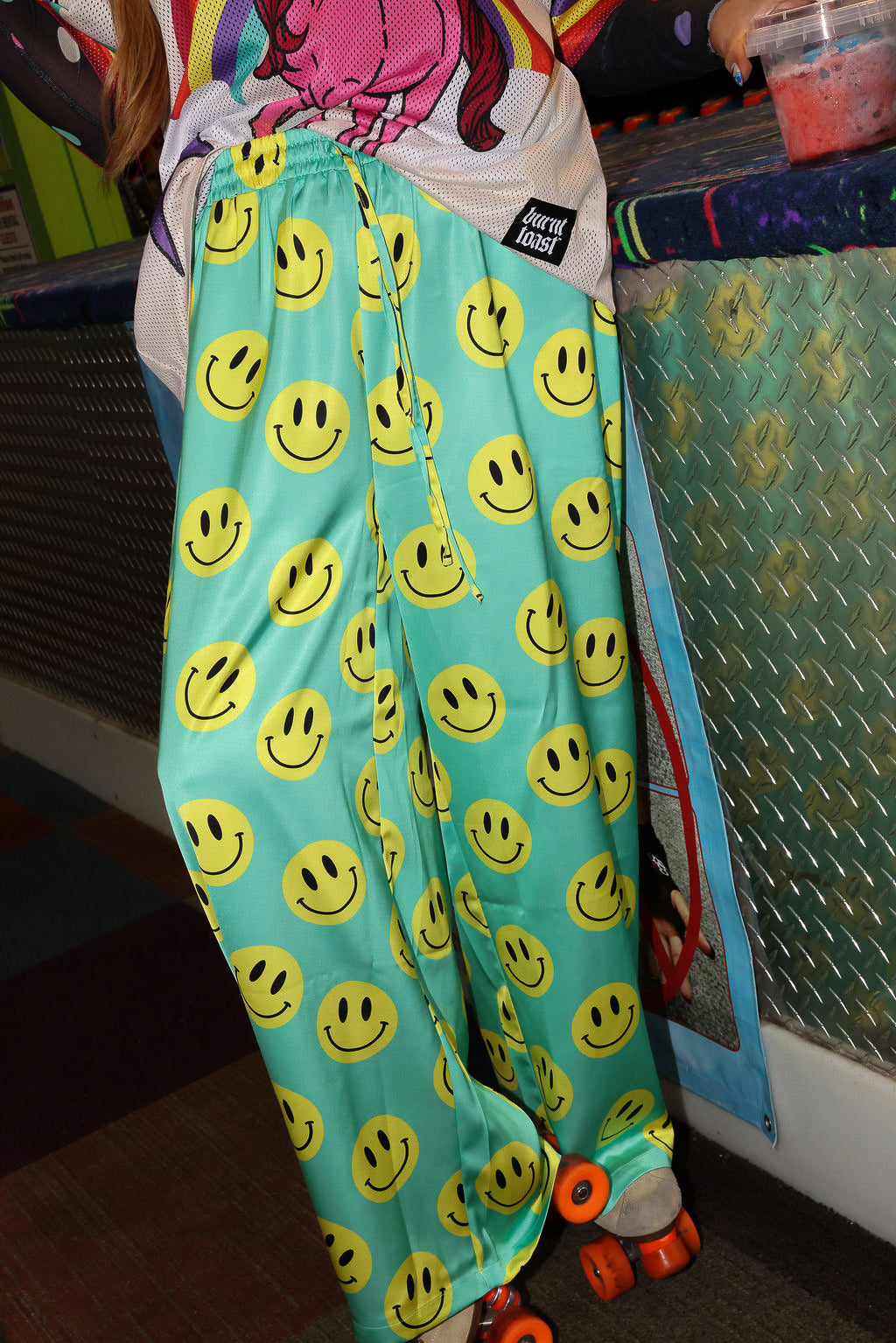 Model wearing seafoam green satin pants with yellow smiley face patterns, and roller skates, close up frontal view