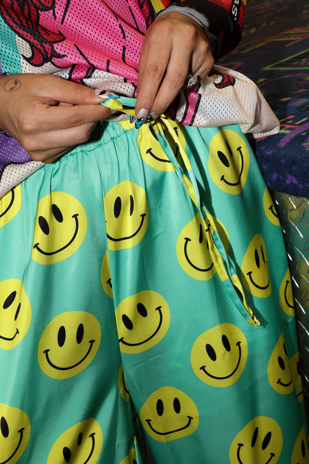 Model wearing seafoam green satin pants with yellow smiley face patterns, holding drawstring waist, close up frontal view