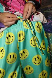Model wearing seafoam green satin pants with yellow smiley face patterns, holding drawstring waist, close up frontal view