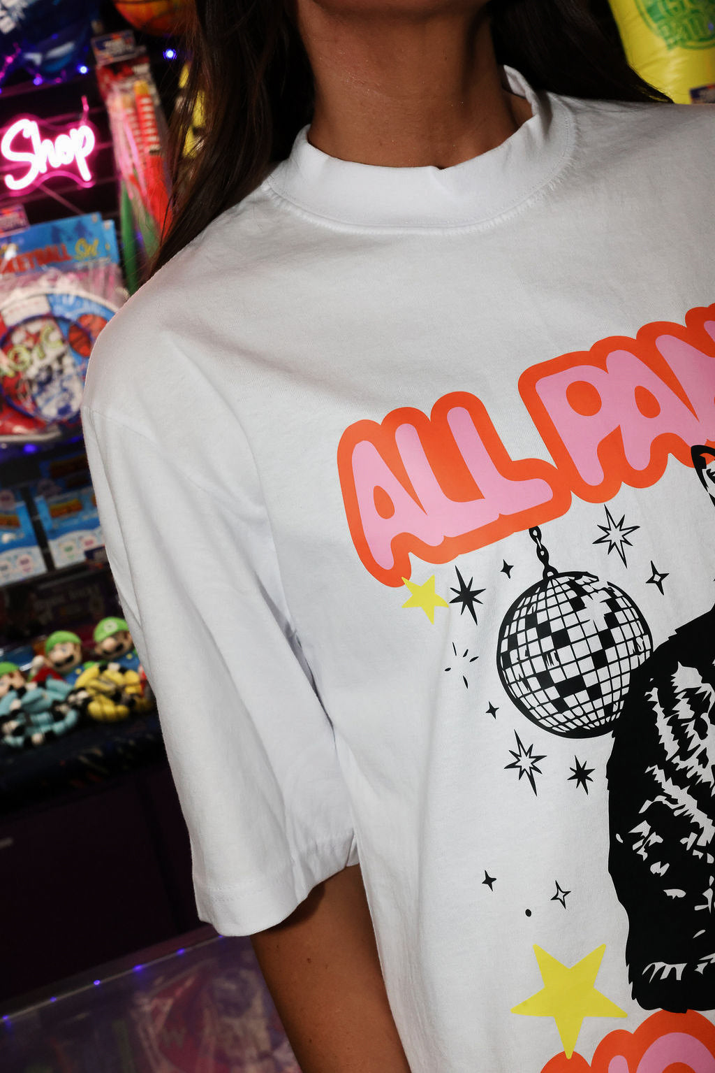 A person wearing a white graphic t-shirt with a colorful 'ALL PANIC NO DISCO' disco-themed print featuring a disco ball and a cat silhouette, set against a blurred arcade background