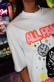 A person wearing a white graphic t-shirt with a colorful 'ALL PANIC NO DISCO' disco-themed print featuring a disco ball and a cat silhouette, set against a blurred arcade background
