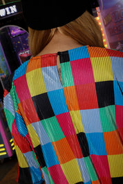 Model wearing a black beanie, a colorful checkered plisse short sleeve top with clasp on the neckline, in front of an arcade machine, close up back view