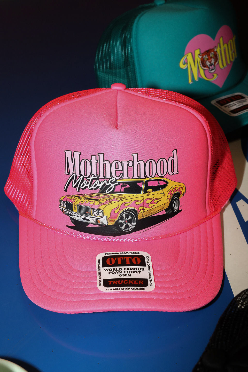 Neon pink trucker hat with 'Motherhood Motors' text and car design on a blue surface, flat lay frontal view