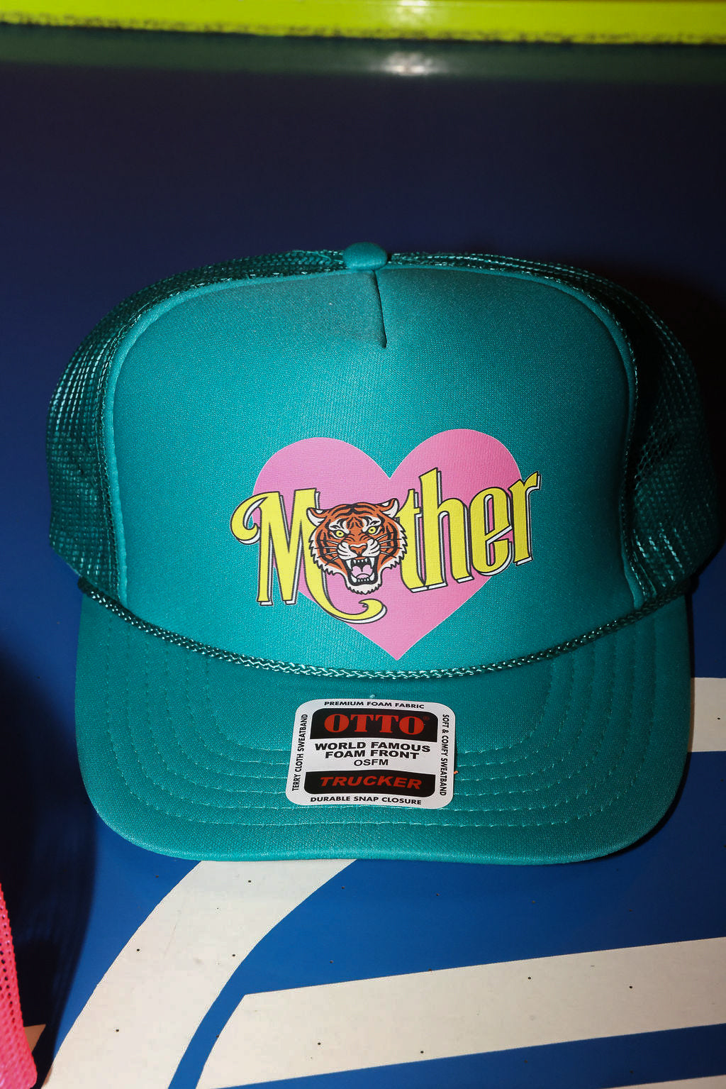 Teal trucker hat with pink heart and yellow  'Mother' text with a tiger, on a blue and white background, flat lay frontal view