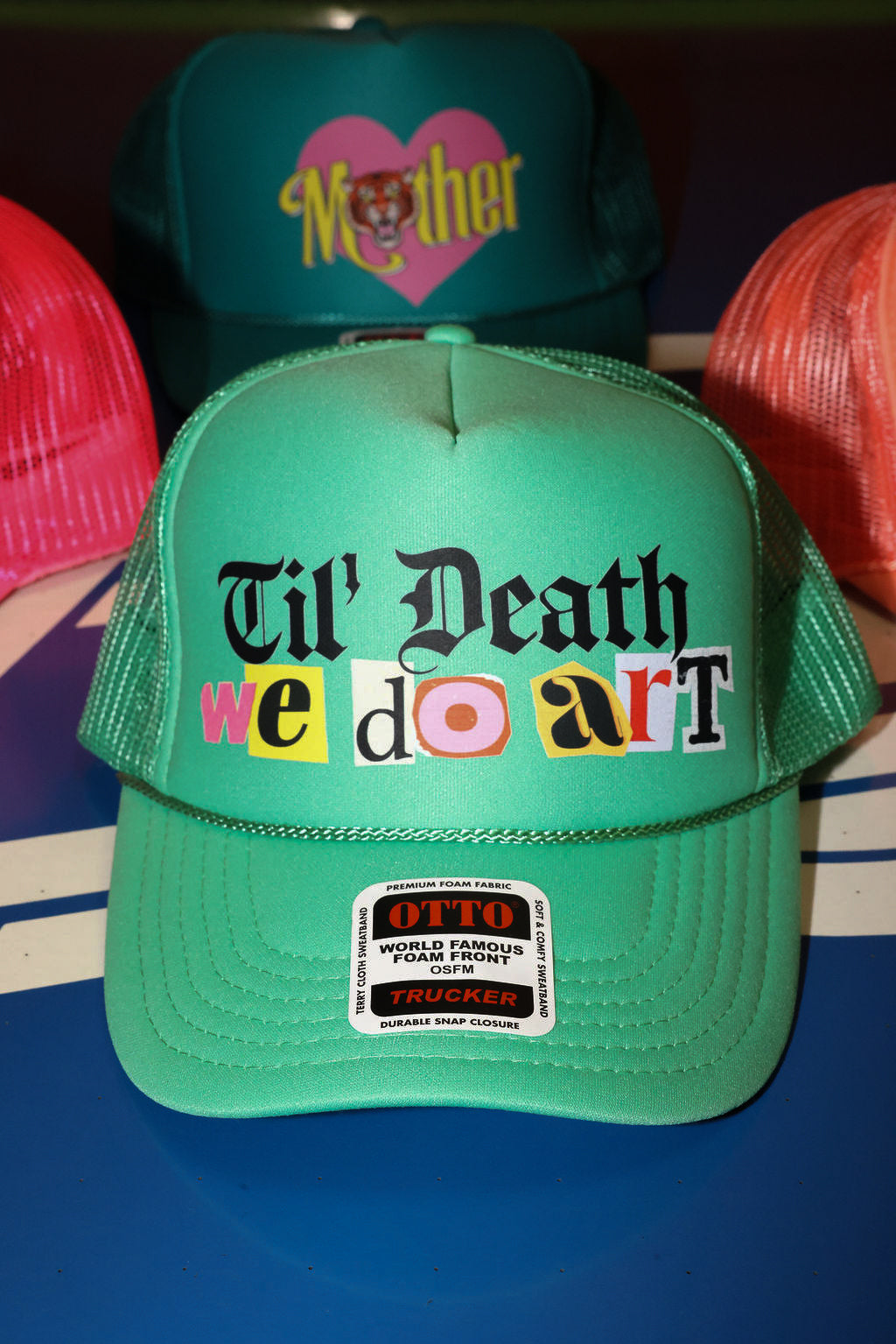 Green trucker hat with 'Til Death We Do Art' text and a trucket hat with 'Mother' logo in the background, flat lay frontal view