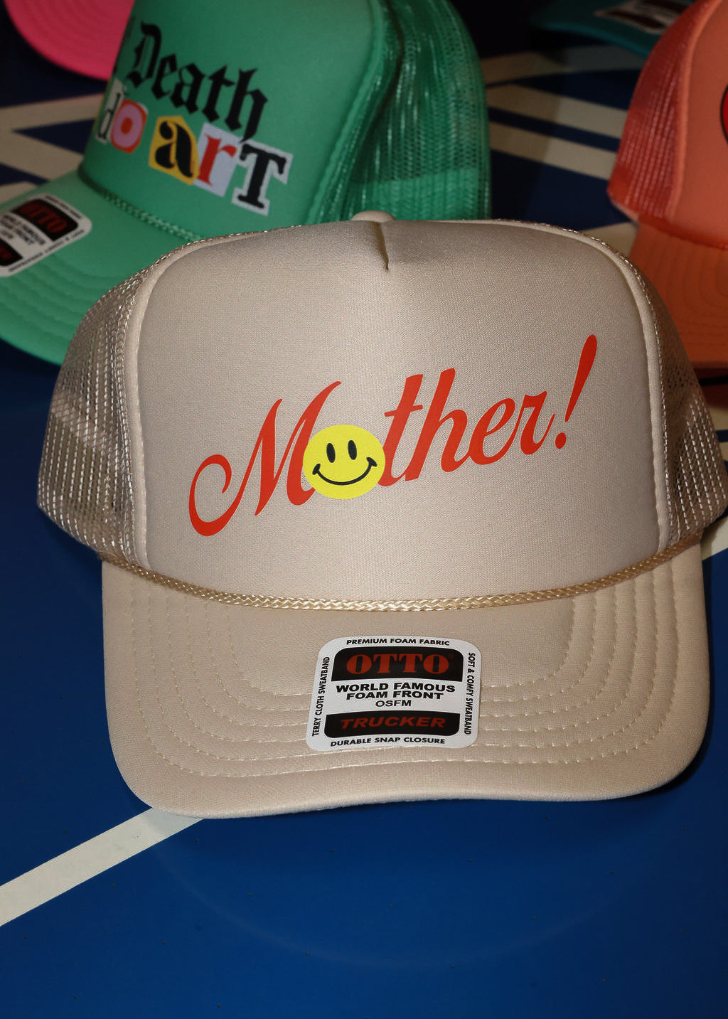 Beige trucket hat with red 'Mother!' text and smiley face on a blue surface, flat lay frontal view
