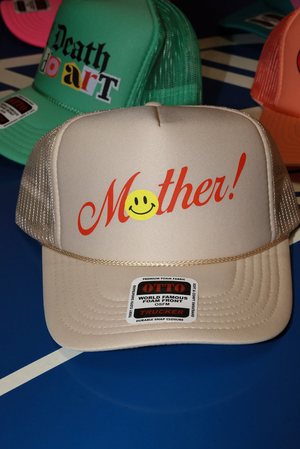 Beige trucket hat with red 'Mother!' text and smiley face on a blue surface, flat lay frontal view