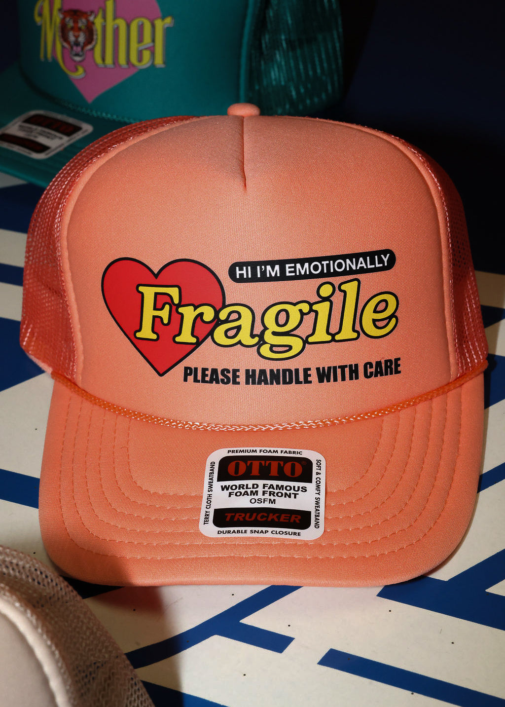 Peach colored trucket hat with 'Hi I'm Emotionally Fragile Please handle With Care' text and heart graphic on a patterned surface, flat lay frontal view