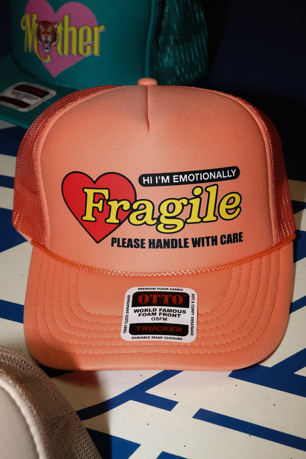 Peach colored trucket hat with 'Hi I'm Emotionally Fragile Please handle With Care' text and heart graphic on a patterned surface, flat lay frontal view