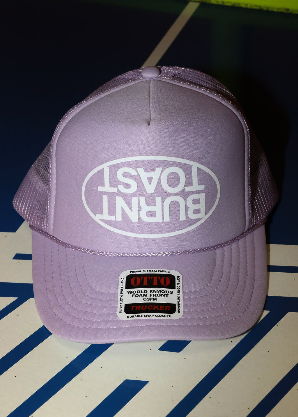 Purple trucker hat with upside down 'Burnt Toast' logo on a striped surface, flat lay frontal view