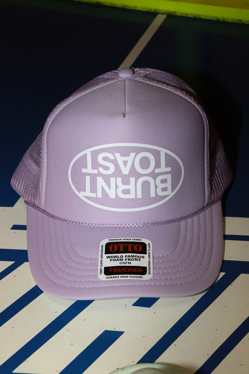 Purple trucker hat with upside down 'Burnt Toast' logo on a striped surface, flat lay frontal view