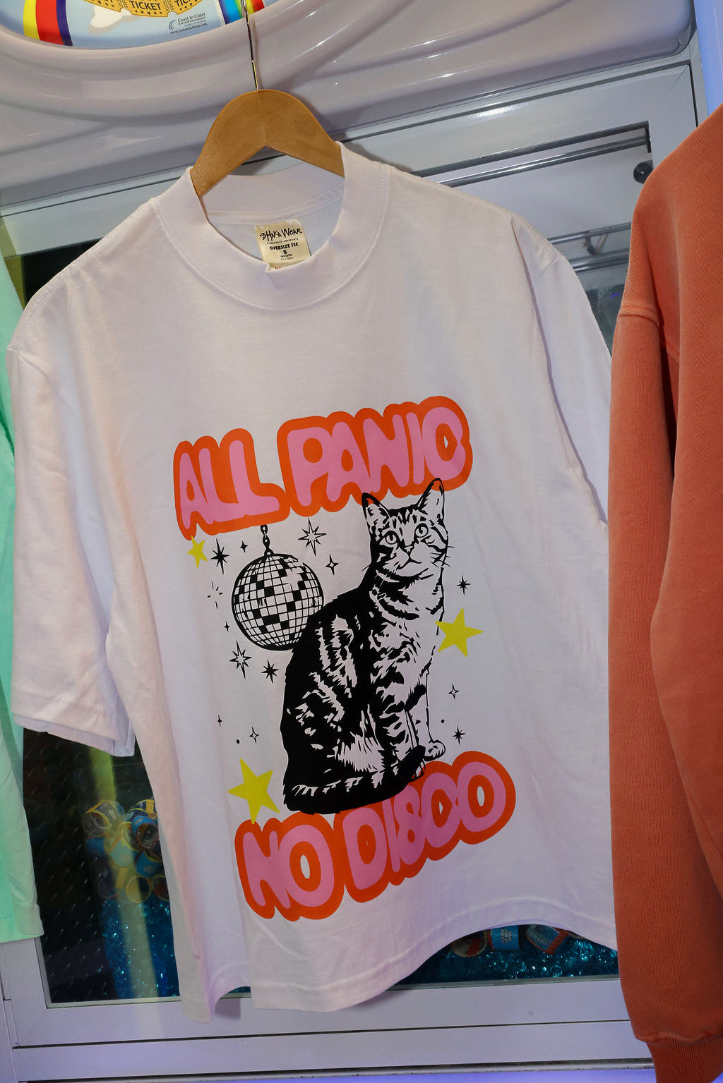 A white graphic t-shirt hanging on a wooden hanger, featuring a black cat illustration with a disco ball and the text 'ALL PANIC NO DISCO' in orange and yellow
