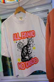 A white graphic t-shirt hanging on a wooden hanger, featuring a black cat illustration with a disco ball and the text 'ALL PANIC NO DISCO' in orange and yellow