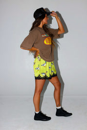 Woman wearing a brown t-shirt with a logo and satin shorts with lace trim and duck design, holding back trucker hat, on a white background, side view