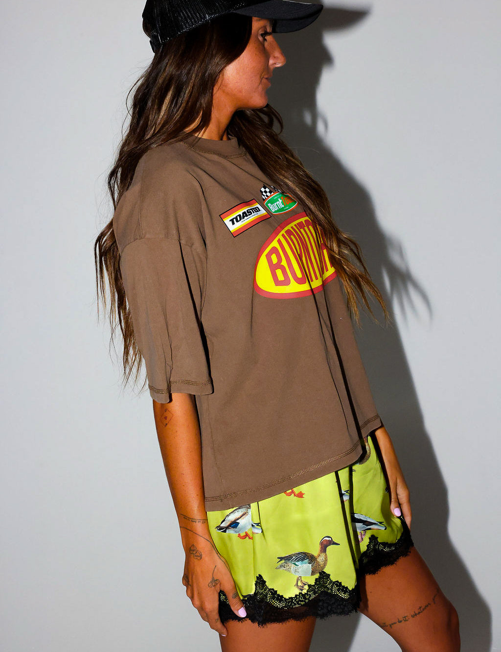 Woman wearing a brown t-shirt with a logo and satin shorts with lace trim and duck design, on a white background, side view