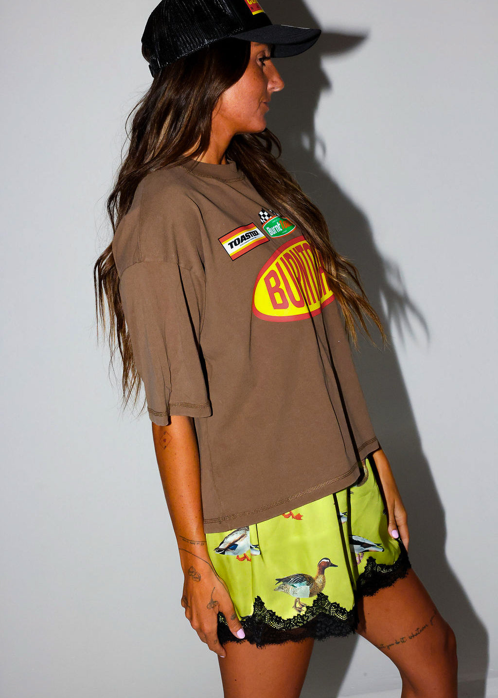 Woman wearing a brown t-shirt with a logo and satin shorts with lace trim and duck design, on a white background, side view