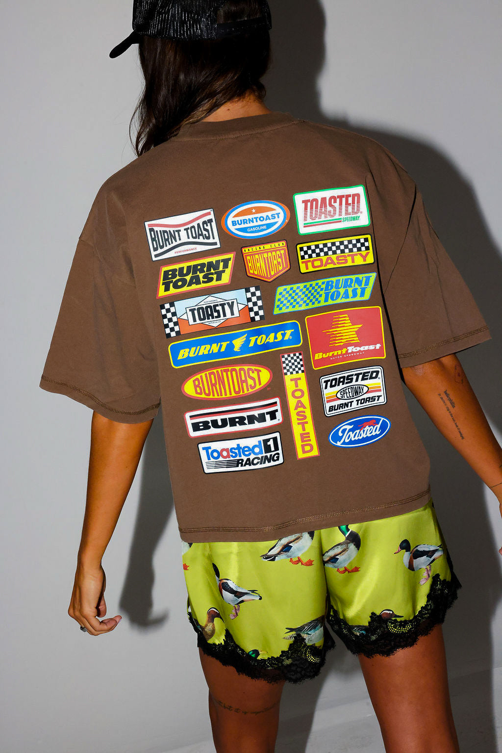 Woman wearing a brown t-shirt with a logo and satin shorts with lace trim and duck design, on a white background, back view