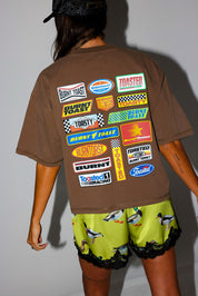 Woman wearing a brown t-shirt with a logo and satin shorts with lace trim and duck design, on a white background, back view